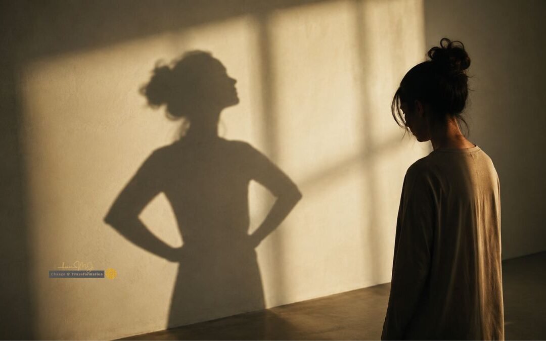 A woman stands quietly with her arms at her sides while her shadow on the wall behind her stands with hands on hips, head lifted, in a confident pose.
